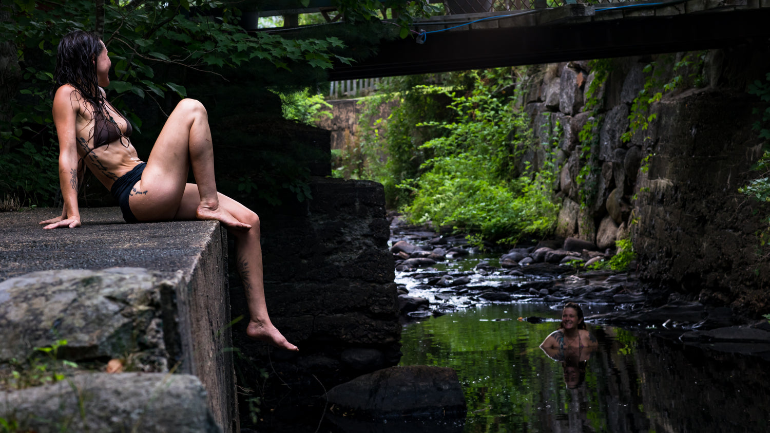 Person sitting on a rock by a stream with another person in the water.