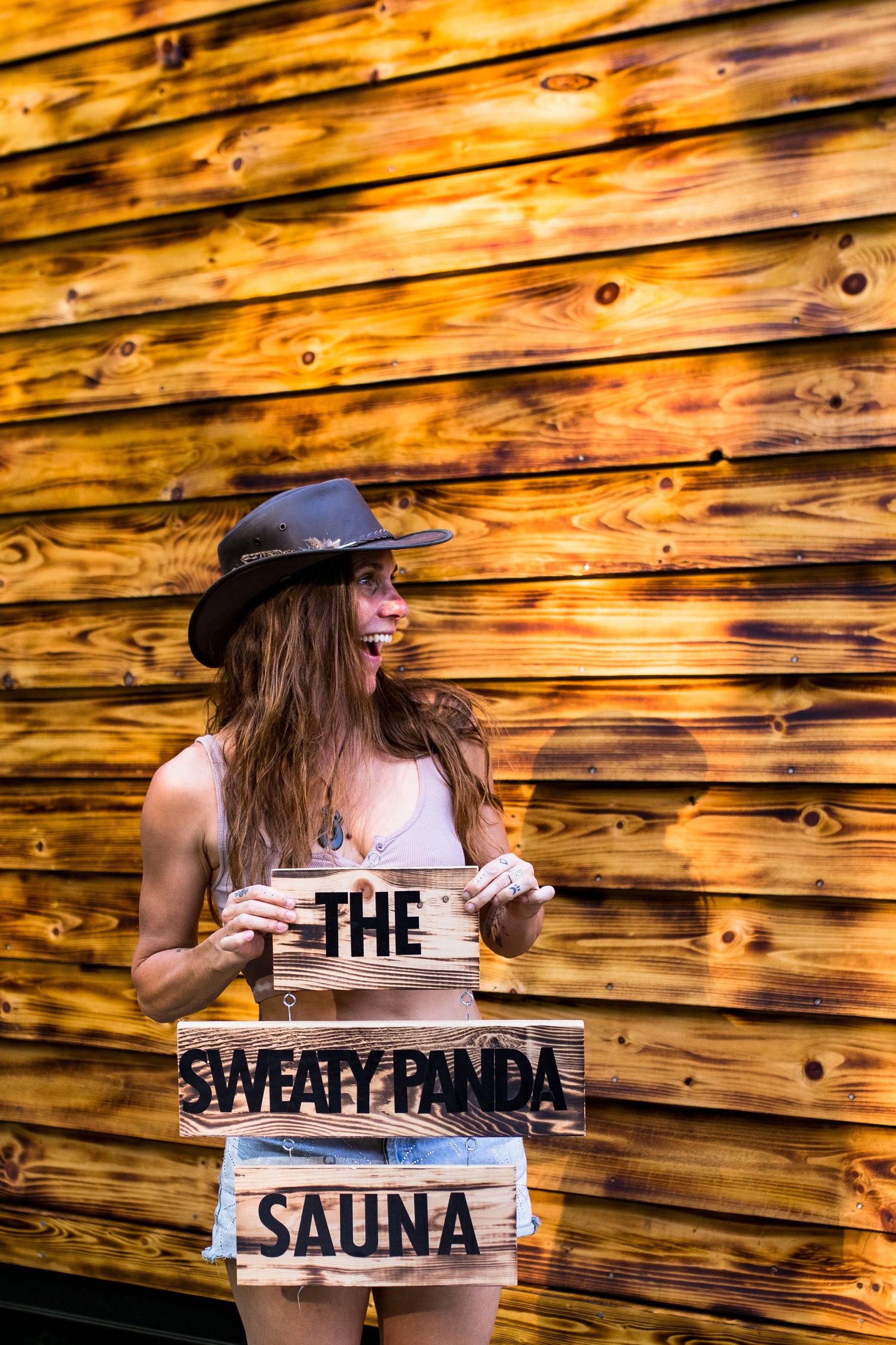 Person holding a sign with 'The Sweaty Panda Sauna' against a wooden wall.